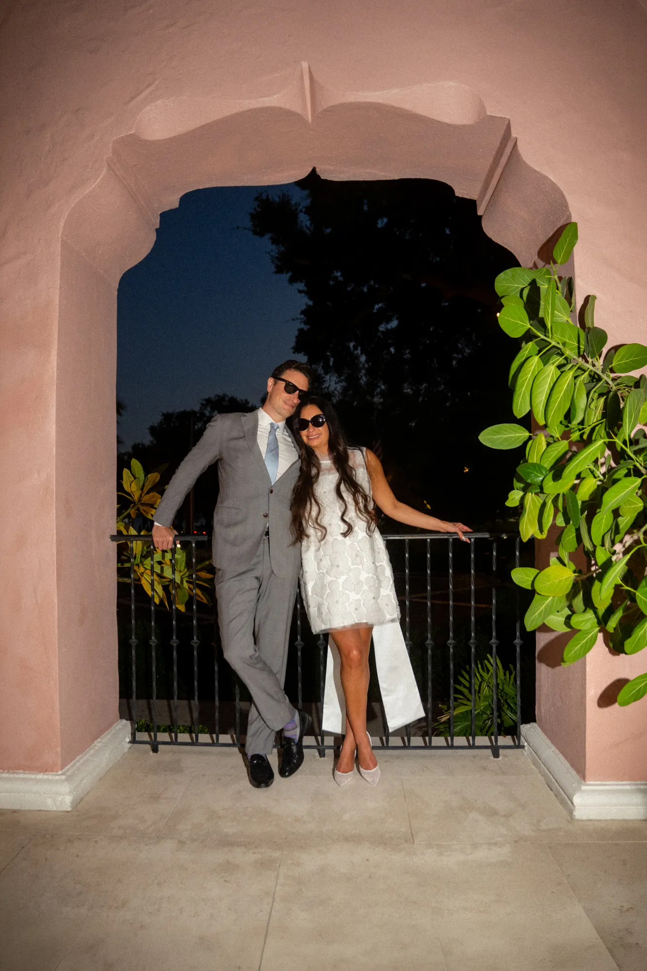 Bride and groom in sunglasses leaning against a pink Mediterranean archway